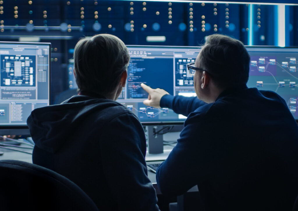 Two Professional IT Programers Discussing Blockchain Data Network Architecture Design and Development Shown on Desktop Computer Display. Working Data Center Technical Department with Server Racks